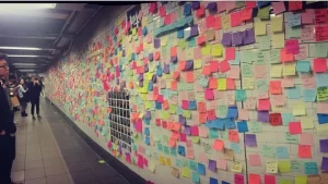 A subway station wall covered with hundreds of colorful sticky notes displaying handwritten messages, with a few people standing nearby reading them.